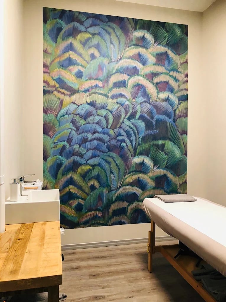 Modern spa or wellness room with a large abstract feather-pattern tapestry in blue and gold tones on the wall, featuring massage or treatment bed and wooden furnishings
