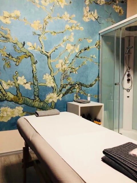Spa treatment room with white massage bed, blue wall featuring Van Gogh almond blossoms mural, glass shower enclosure, and white storage shelf